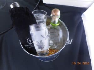 a Corona beer bucket filled with crinkle paper. Also holding a bottle of tequila, and two glasses, and a bag containing a cocktail shaker.