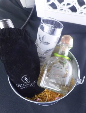 a Corona beer bucket filled with crinkle paper. Also holding a bottle of tequila, and two glasses stacked, and a bag containing a cocktail shaker.