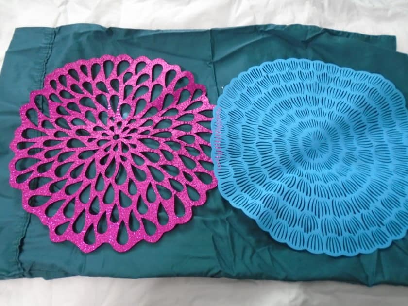 two placemats placed on top of a peacock green pillowcase. Placemates are floral designs. one is foam with pink glitter. the other is a blue vinyl material.