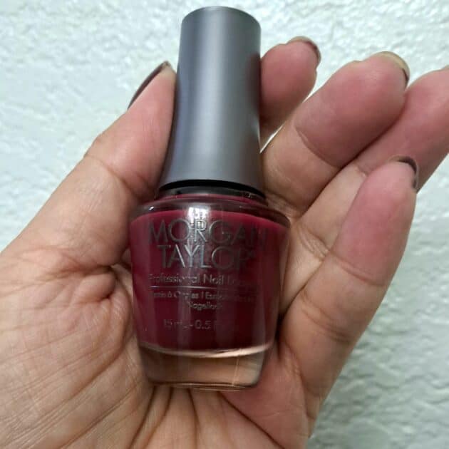 Morgan Taylor nail poish bottle in (V) my hand. Nail polish shade is deep red. Botle has a gray lid.